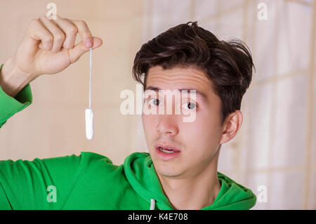 Portrait of handsome man with a surprised face holding a menstruation cotton tampon, wearing a green hoodie in a blurred background Stock Photo