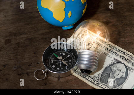 Navigation compass, lit light bulb, and a world globe on top of a US one dollar bill, with a wooden background Stock Photo