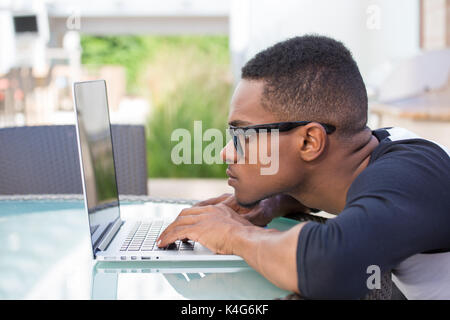 Closeup portrait, young nerdy man in big black glasses surfing the web on personal silver laptop, isolated outside outdoors background Stock Photo