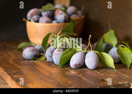 Fresh plums with green leaves in wooden pot on the dark wooden table. Shallow depth of field. Toned. Stock Photo