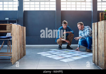 Two focused young work colleagues brainstorming together while crouching over papers spread out in a square on the floor of a modern office Stock Photo