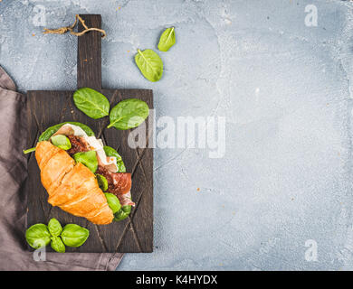 Croissant sandwich with smoked meat Prosciutto di Parma, sun dried tomatoes, fresh spinach and basil on dark wooden board over stone textured grey background Stock Photo