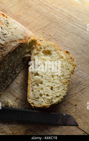 Overhead shot of freshly baked loaf of bread. First slice cut and lying face up on wooden chopping board with knife blade. Stock Photo