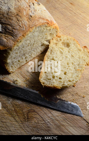 Freshly baked loaf of bread, shot from above. First slice cut and lying face up on wooden chopping board with knife blade. Stock Photo