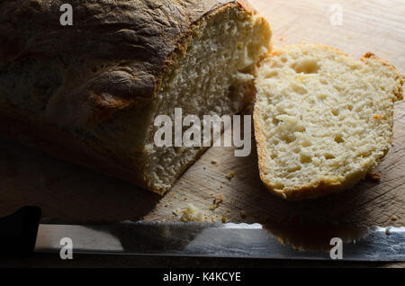 Freshly baked loaf of crusty bread with first slice cut and lying face up on wooden chopping board alongside serrated knife. Stock Photo