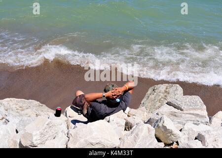 Zion, Illinois, USA. A lone man sitting among the rocks well above a beach at Illinois Beach State Park. Stock Photo