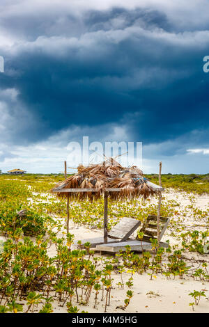 Broken wooden sunloungers and a derelict palm leaf covered parasol on a beach in south Barbuda, Antigua & Barbuda, West Indies under stormy grey sky Stock Photo