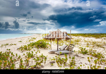 Broken wooden sunloungers and a derelict palm leaf covered parasol on a beach in south Barbuda, Antigua & Barbuda, West Indies under stormy grey sky Stock Photo