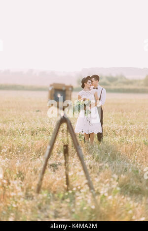 The lovely photo of the newlyweds hugging behind the blurred view of the old vintage camera. The groom is hugging the bride back. Stock Photo