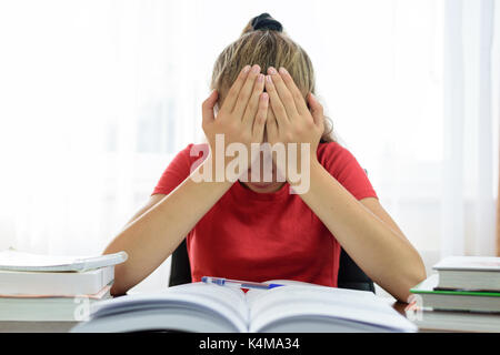 Schoolgirl holds her head in her hands over a pile of books on the desk and is tired and frustrated with problems at school. Stock Photo