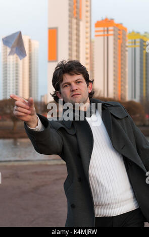 Paper airplane launching in park, free space, Man in grey coat and white sweater holding handmade paper airplane in hand on high-rise building background. Open-air game, rest, leisure, pastime concept Stock Photo