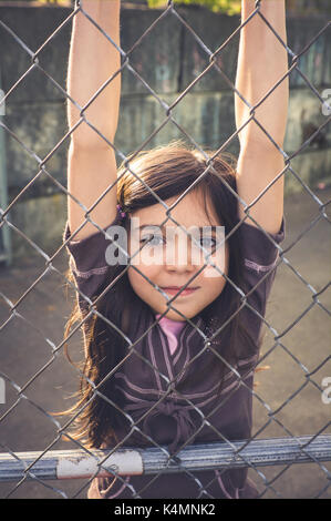 A young girl hanging on a chainlink fence, playing outside in an urban setting. Stock Photo