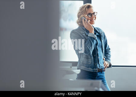 Smiling woman standing in office and talking on smartphone. Young woman using mobile phone in the office. Stock Photo