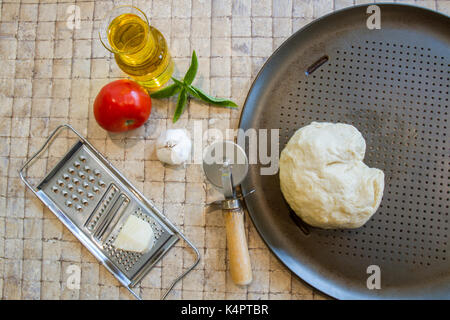Fresh basil, whole ripe tomato, olive oil and cheese with fresh dough on baking sheet and pizza cutter on kitchen tile. Stock Photo