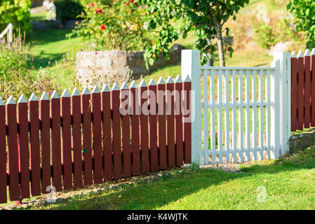 Red picket fence with white gate and garden in background. Stock Photo