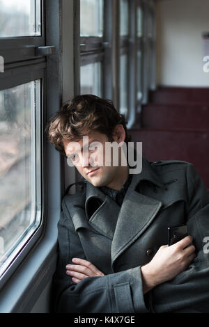 Traveling by train. Sad man traveling by train, looking through the window and thinking about unrequited love squeezing the phone in his hand. alone in an empty train wagon. Vertical. Stock Photo