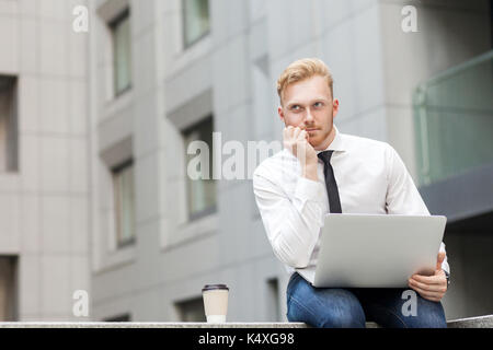 Working in outdoor, sits on the ground. Young adult ginger bearded businessman , work in computer and dreaming. Outdoor shot Stock Photo