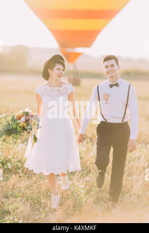 The close-up vertical portrait of the happy newlyweds holding hands and walking in the field during the sunset. The couple is dressed in the vintage style. Stock Photo