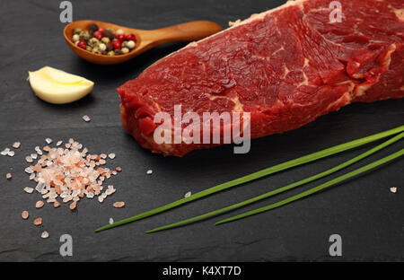 One raw beefsteak red meat cut, spices, peppercorn in wooden scoop, green spring chive, garlic and Himalayan salt on black slate board, close up, high Stock Photo
