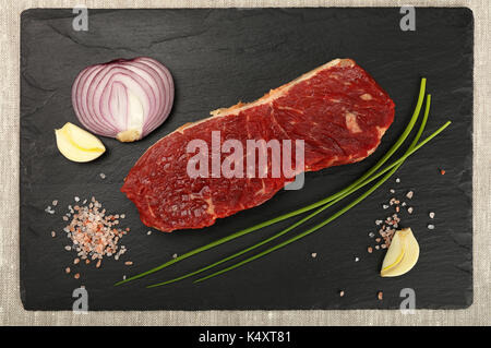 One raw beefsteak red meat cut, spices, peppercorn in wooden scoop, green spring chive, onion, garlic and Himalayan salt on black slate board, close u Stock Photo