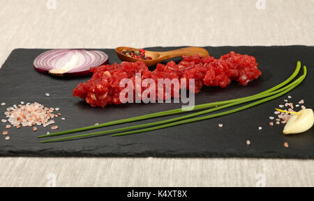 Raw minced beef red meat cutlet, spices, peppercorn in wooden scoop, spring green chive, onion, garlic cloves and Himalayan salt on black slate board, Stock Photo