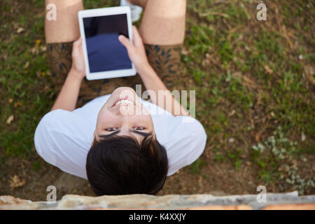 Upside down portrait of young happy man sitting on grass holding tablet Stock Photo