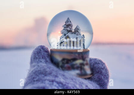 Girl in knitted mittens holding glass ball with firtrees, house and artificial snow with winter landscape at the  background Stock Photo