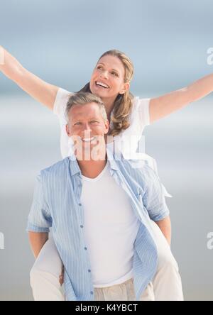 Digital composite of Middle aged couple piggy back against blurry beach Stock Photo