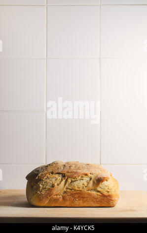 A single, whole, crusty loaf of bread on a wooden chopping board against white tiled background with copy space above. Stock Photo