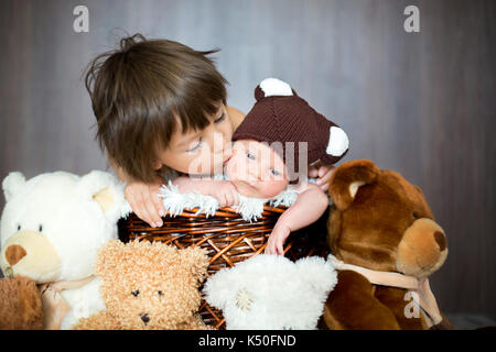 Cute newborn baby boy in basket with teddy bear hat,older brother hugging him and giving him a kiss, teddy bear toys around them Stock Photo