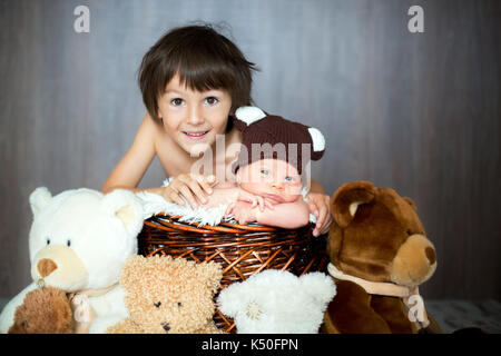 Cute newborn baby boy in basket with teddy bear hat,older brother hugging him and giving him a kiss, teddy bear toys around them Stock Photo