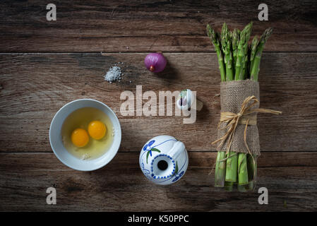 Bunch of fresh asparagus, with eggs, onion and salt on rustic wooden background. Ingredients ready to make a meal. Top view. Stock Photo
