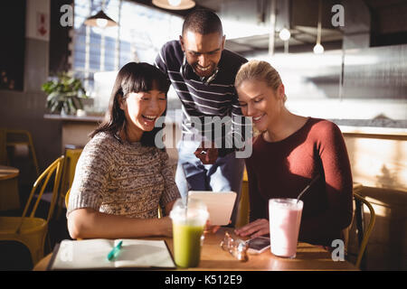 Smiling man pointing at digital tablet while standing amidst female friends in coffee shop Stock Photo