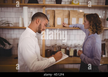 Side view of male owner writing on clipboard while waitress arranging products at coffee shop Stock Photo