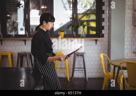 Side view of young waitress reading menu while leaning on table at coffee shop Stock Photo
