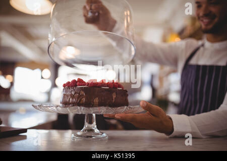 Close up of young waiter holding glass lid over cake on cakestand at counter in coffee shop Stock Photo