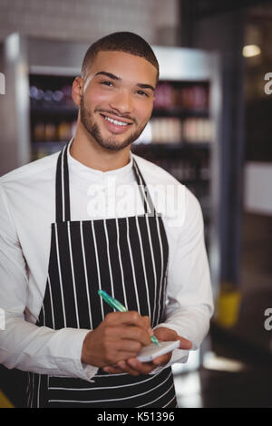 Portrait of smiling young waiter standing with notepad and pen at coffee shop Stock Photo
