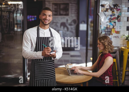 Portrait of handsome waiter standing with notepad by female customer using laptop at coffee shop Stock Photo