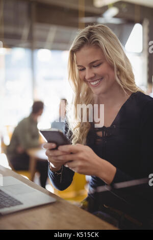 Smiling young blond woman texting through mobile phone while sitting at counter in coffee shop Stock Photo