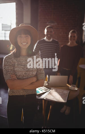 Portrait of smiling young woman wearing hat standing with arms crossed against colleagues at coffee shop Stock Photo