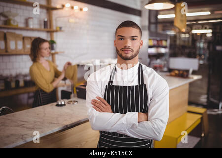 Portrait of confident waiter standing with arms crossed against waitress working at counter in coffee shop Stock Photo