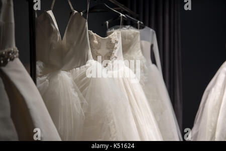 Selection of handmade white wedding gowns hanging on a rail in a darkened room partially illuminated by a light Stock Photo