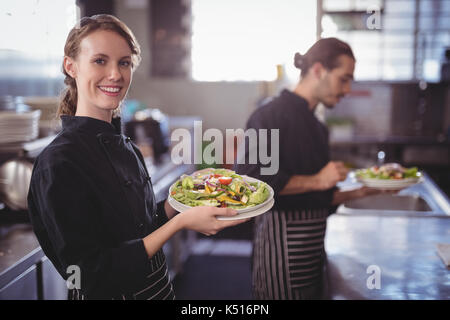 Portrait of smiling young waitress holding fresh salad against waiter at commercial kitchen in coffee shop Stock Photo