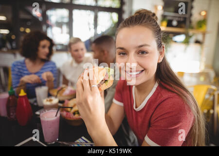 Close up portrait of smiling beautiful young woman eating burger at coffee shop Stock Photo