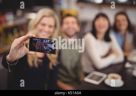 Woman taking selfie with friends from mobile phone while sitting at table in coffee shop Stock Photo