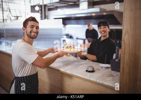 Portrait of smiling female chef and waiter holding plate at counter in coffee shop Stock Photo