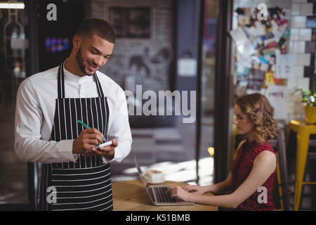 Smiling young waiter writing in notepad while standing against female customer at coffee shop Stock Photo