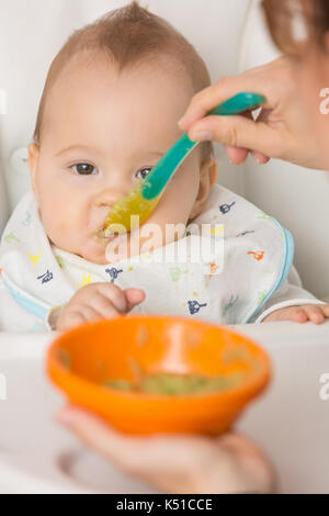 Mother feeding baby girl with mashed avocado and green vegetable in an orange plastic bowl, food on a plastic spoon; concept of family life, healthy e Stock Photo