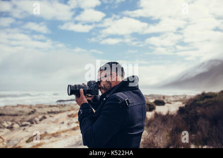 Side view shot of young photographer photographing outdoors. Man taking a photos with dslr camera outdoors. Photographer taking travel nature photogra Stock Photo