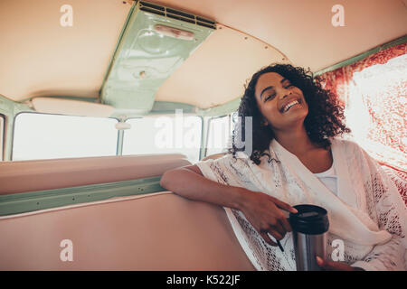 Young woman sitting in backseat of van with coffee and smiling. African woman enjoying on road trip. Stock Photo